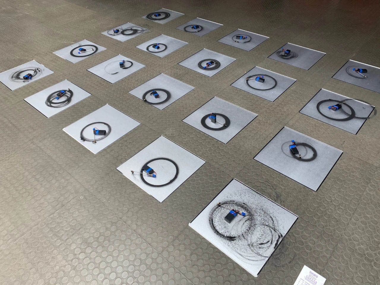 Twenty square boards laid on a grey floor, each with a small motor that has drawn black charcoal circles