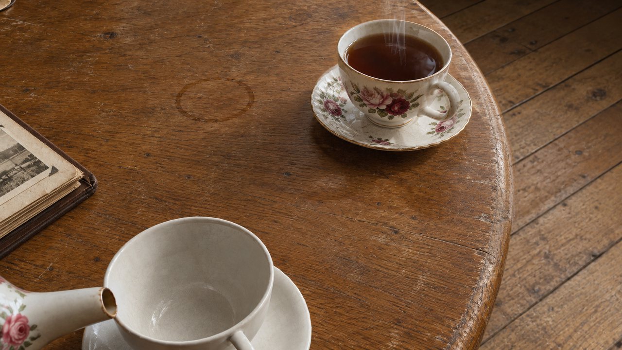 The same view as a finished picture: a steaming cup of tea, an empty cup, a teapot spout and an old album on a worn wooden table.