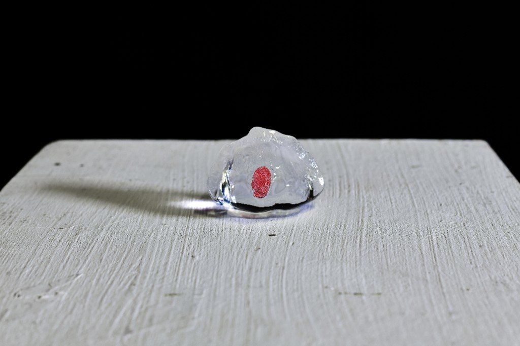 A lump of ice on a white-painted plinth, a red fingerprint-shaped mark inside it and a thin pool of meltwater at its base.